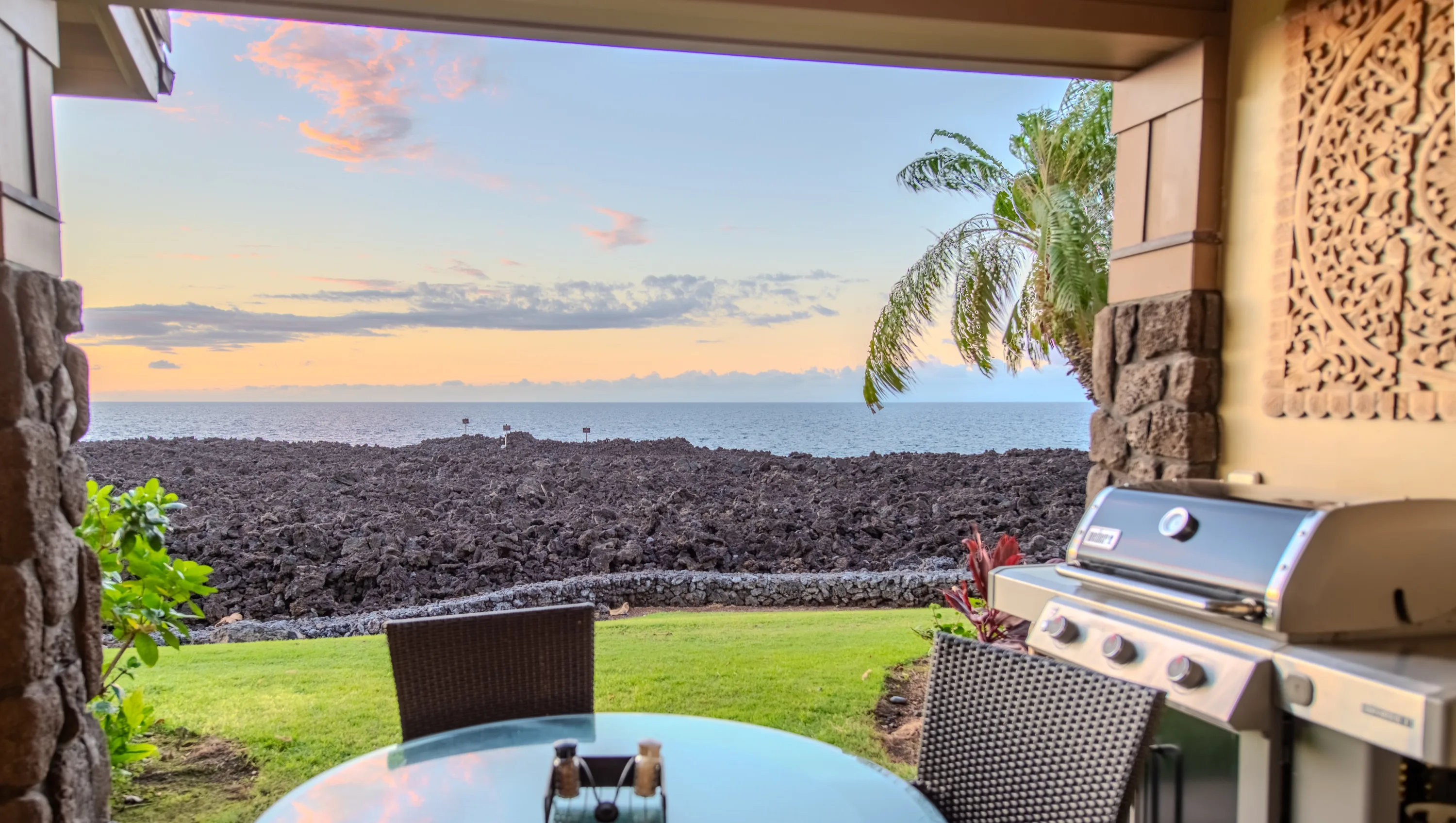 Oceanfront lanai at the Makai Suite with a dining table and grill overlooking the lava-rock shoreline and the Pacific at dusk