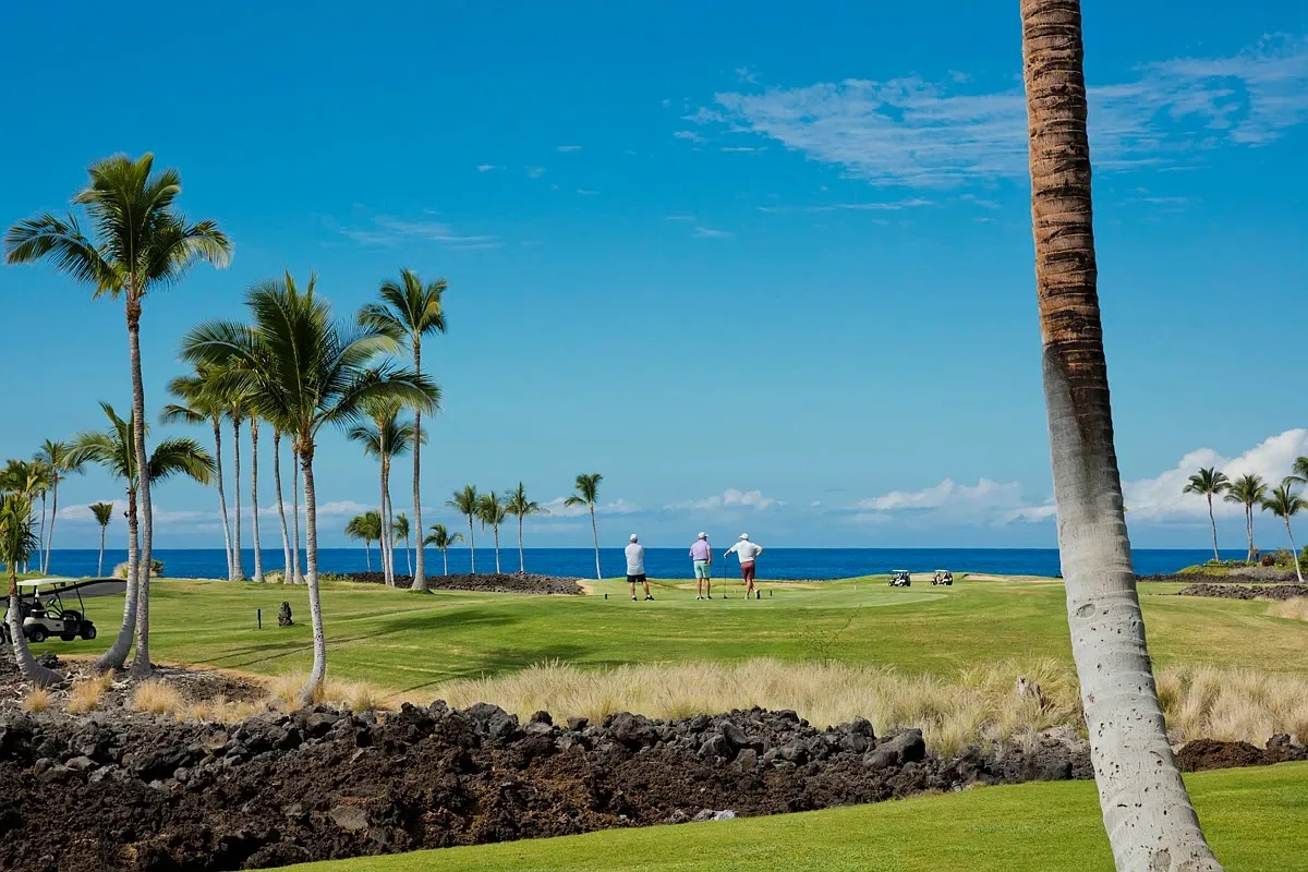 Golfers on the Waikoloa Beach golf course fairway below the Ānuenue Suite, framed by palm trees and lava rock with the Pacific beyond