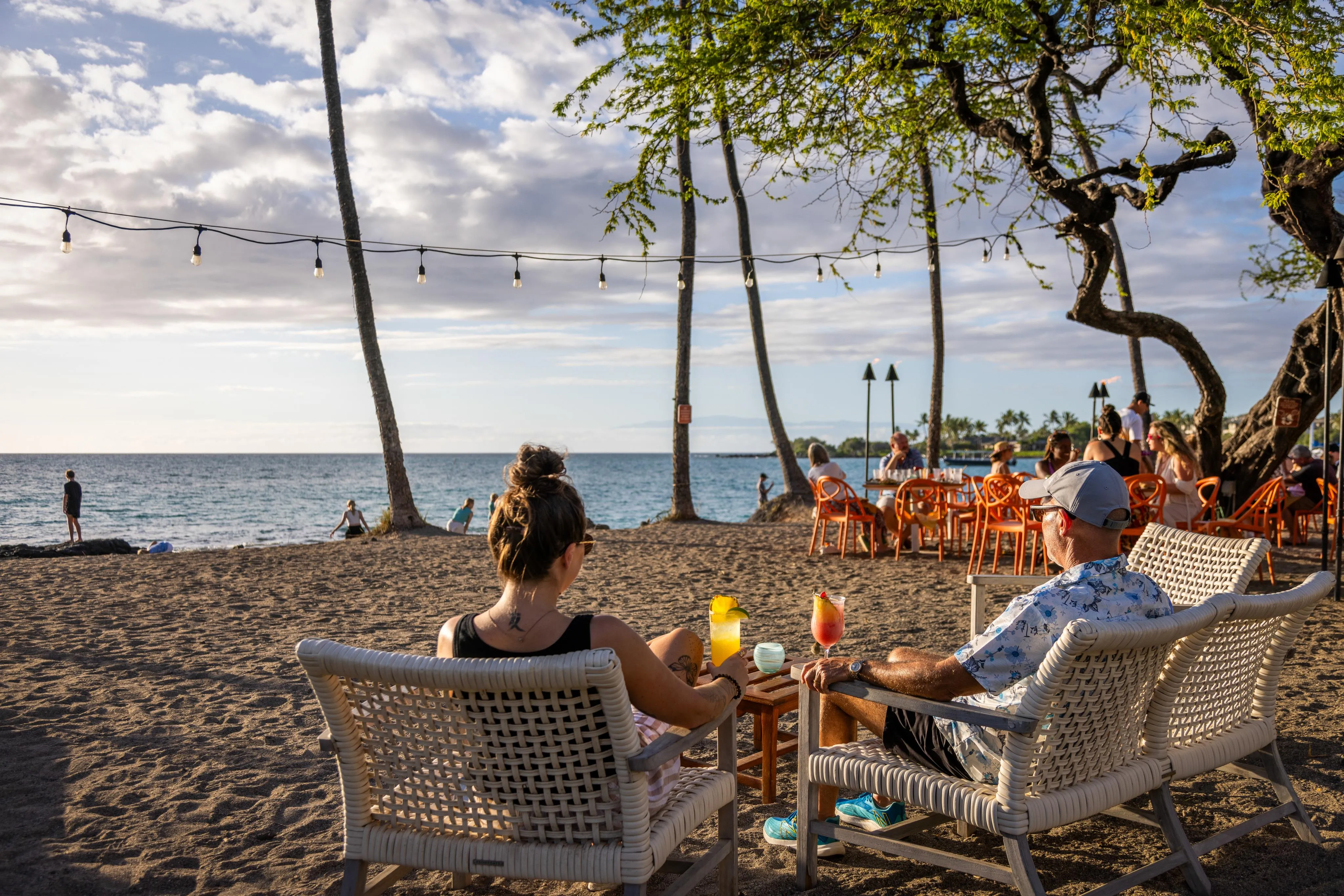 A couple relaxing in beach chairs with cocktails and string lights overhead