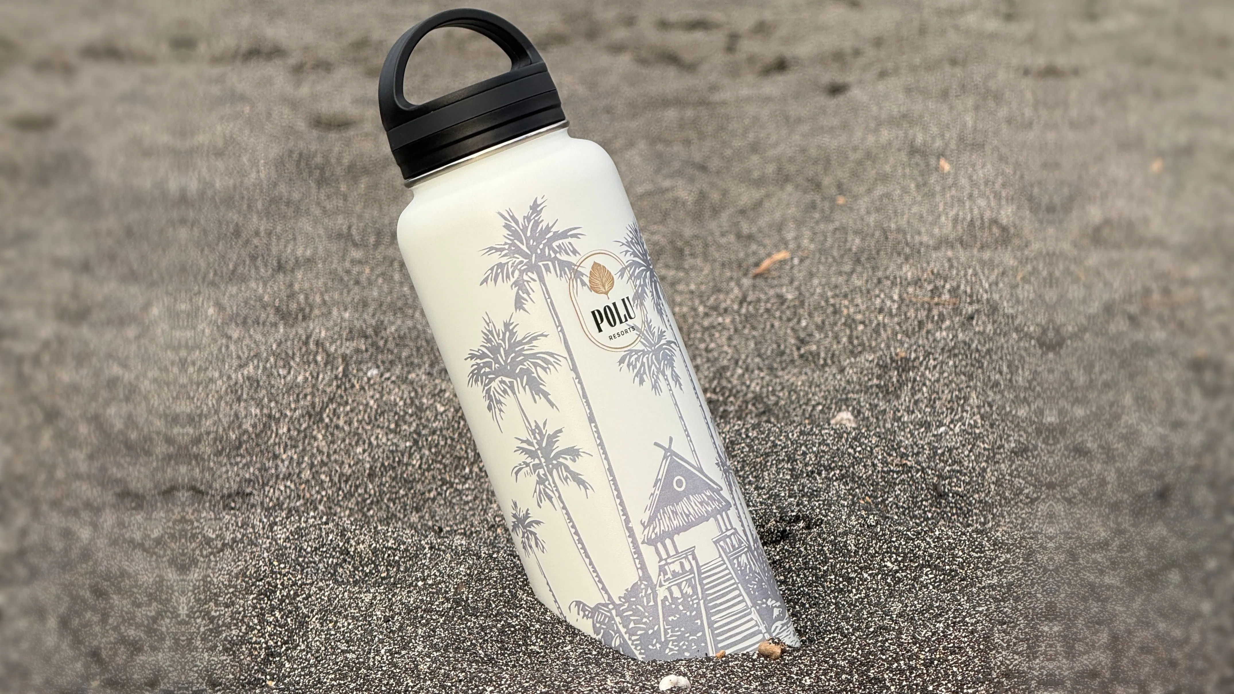 A Polu Resorts insulated reusable water bottle standing upright in black sand on a Hawai'i Island beach