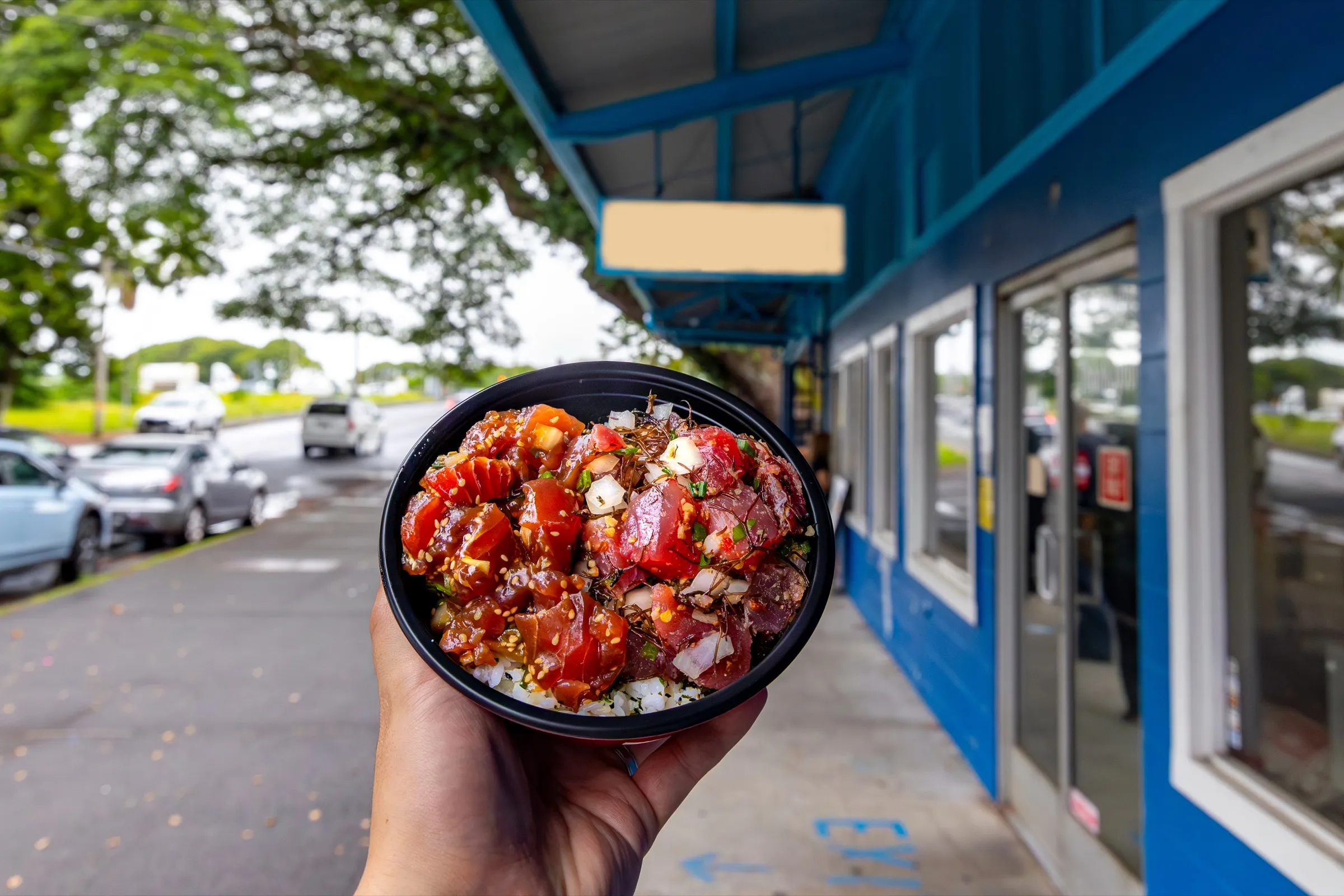 A hand holding a fresh poke bowl outside a local roadside shop